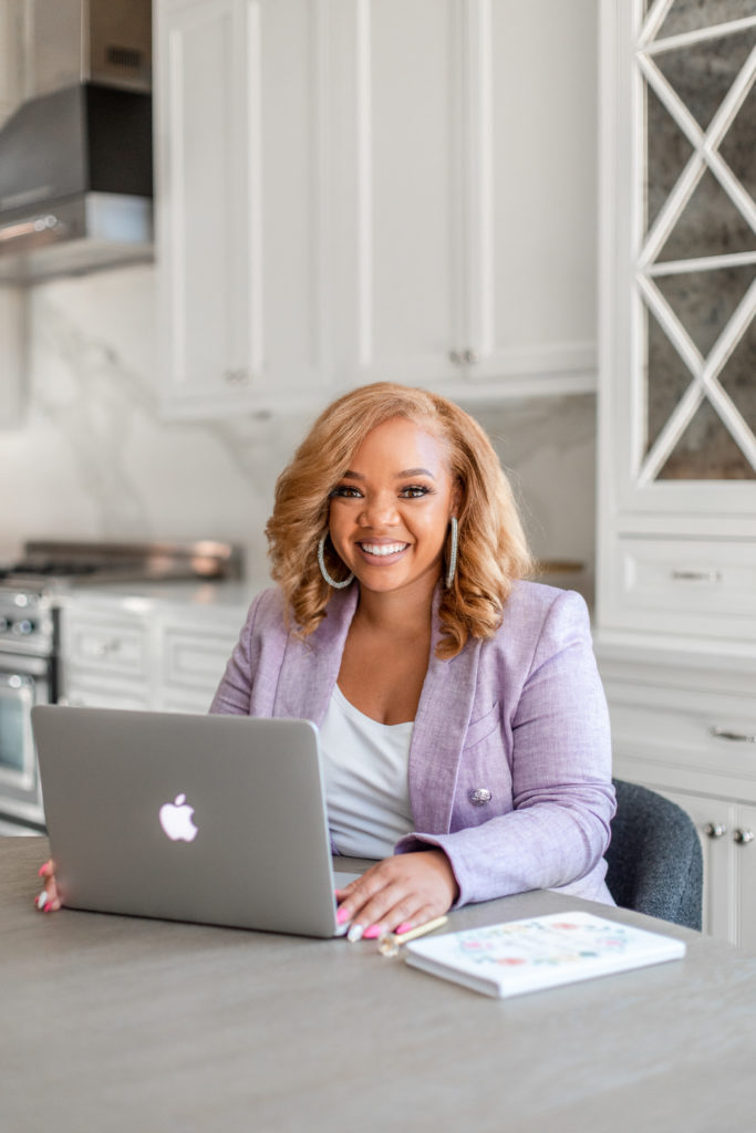 Candace Junée, a beautiful black woman with blonde hair is sitting at a desk in a kitchen with a laptop in front of her. She's in a professional but fashionable purple suit with a big smile on her face. This post is sharing how to make 6-Figures from home.