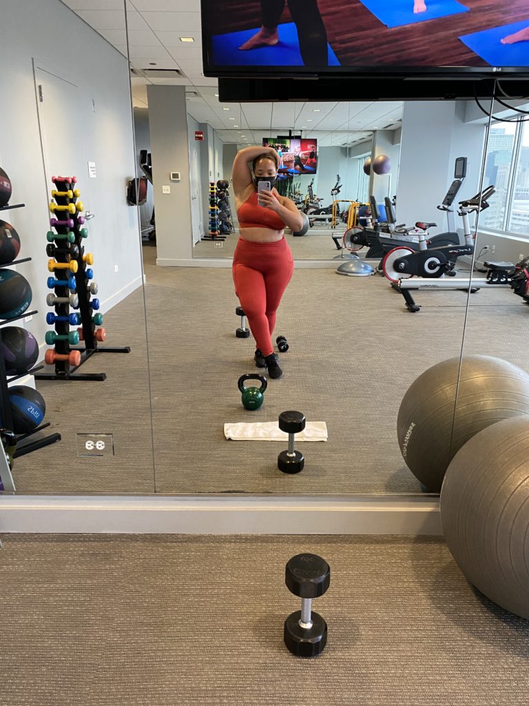 Black woman at the gym taking a picture surrounded by weights and workout equipment.